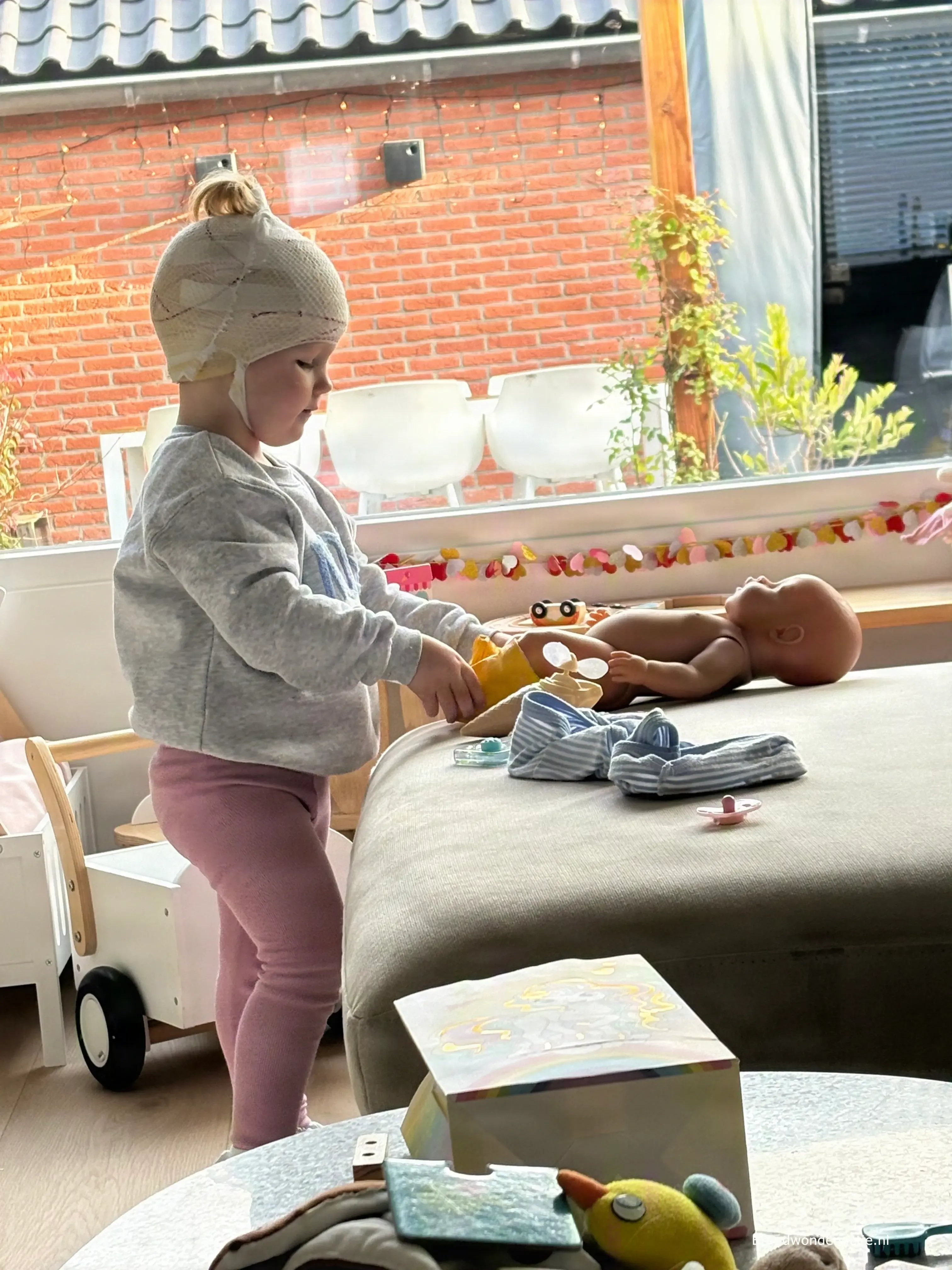 Guusje speelt thuis met een knuffelpop, met nog een verband om haar hoofd.