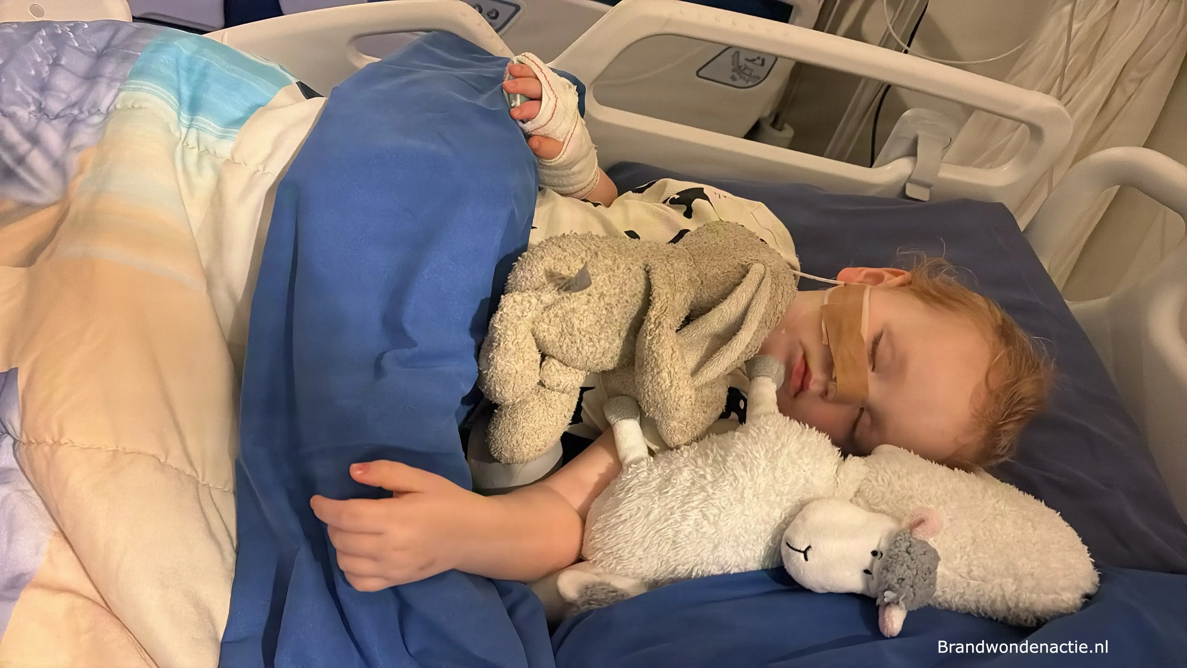Guusje slaapt in het ziekenhuisbed, met haar hand in verband en een knuffel dichtbij.