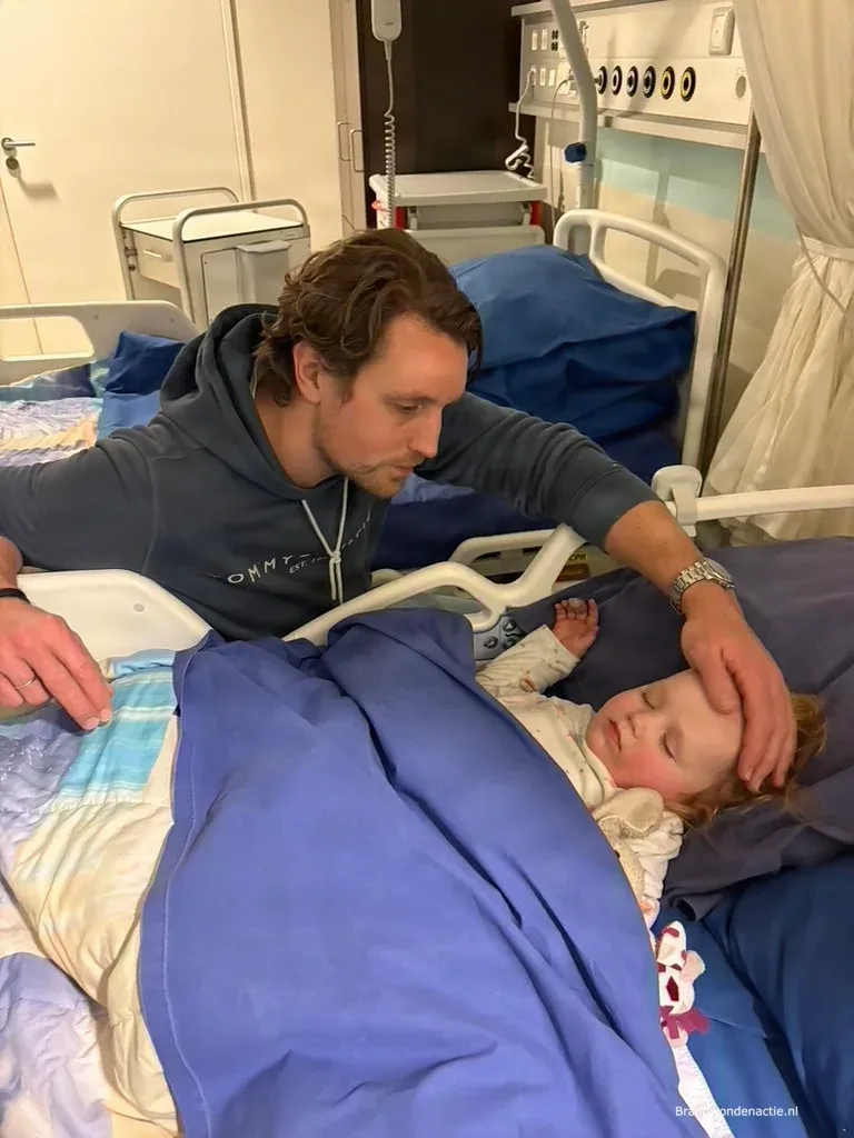 Guusjes vader buigt zich over haar heen en houdt zijn hand op haar hoofd terwijl ze in het ziekenhuisbed ligt.
