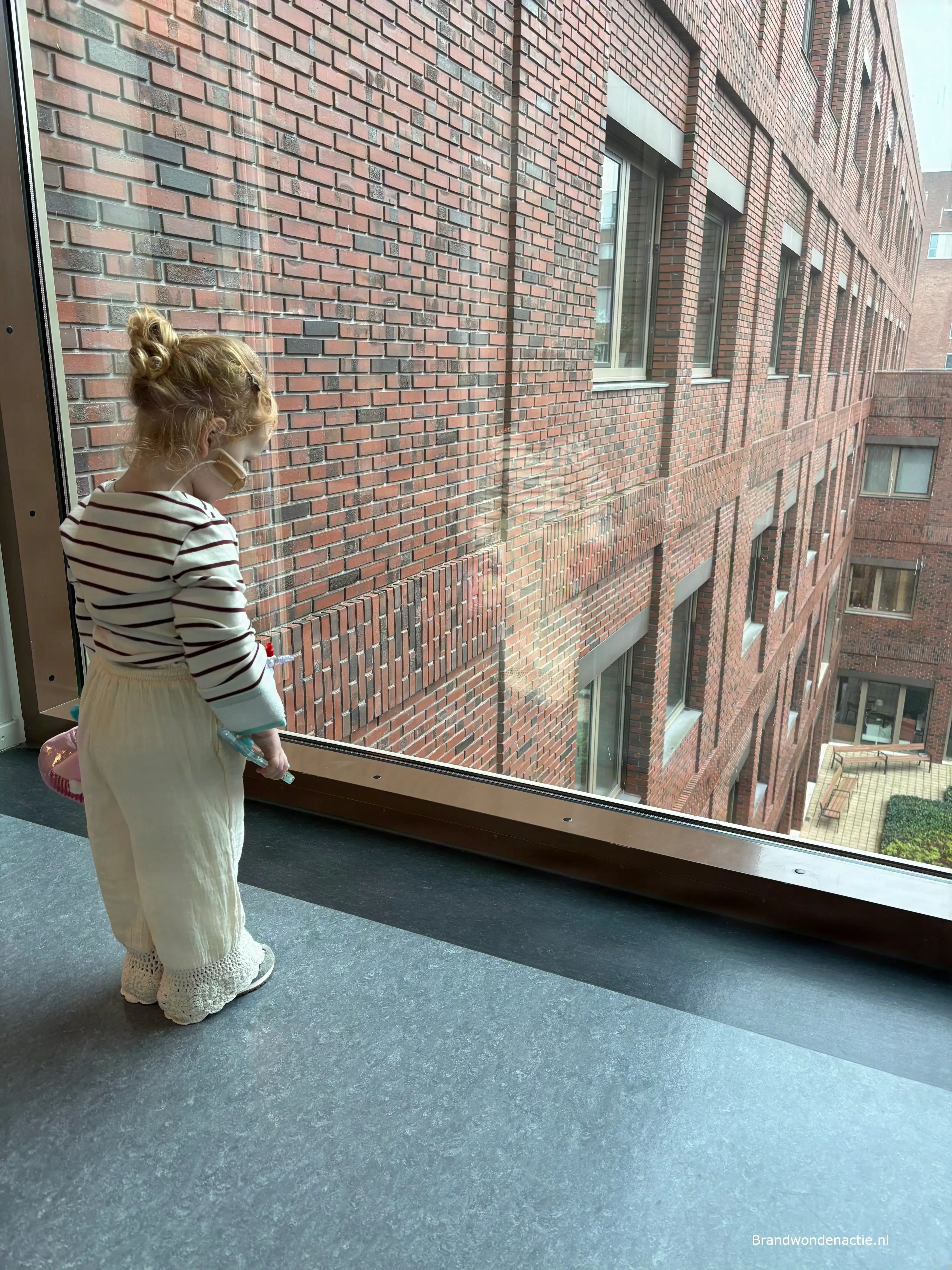 Guusje kijkt door een groot raam naar buiten, vanuit het ziekenhuis.